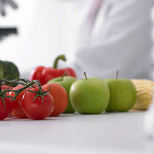 Close-up section of an image showing fruits and vegetables on a table next to a scientist with microscope.