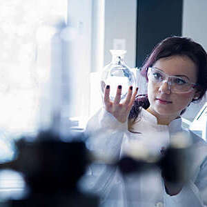 Female biochemistry scientist in lab coat and protective glasses holds up a round flask to study its content.