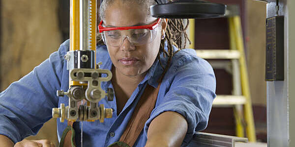 Female technician wearing protection glasses and overall operates a bandsaw machine.