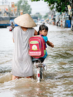 Mother with Vietnamese straw had, pictured from behind, wheels her young daughter on a bicycle through flood waters.